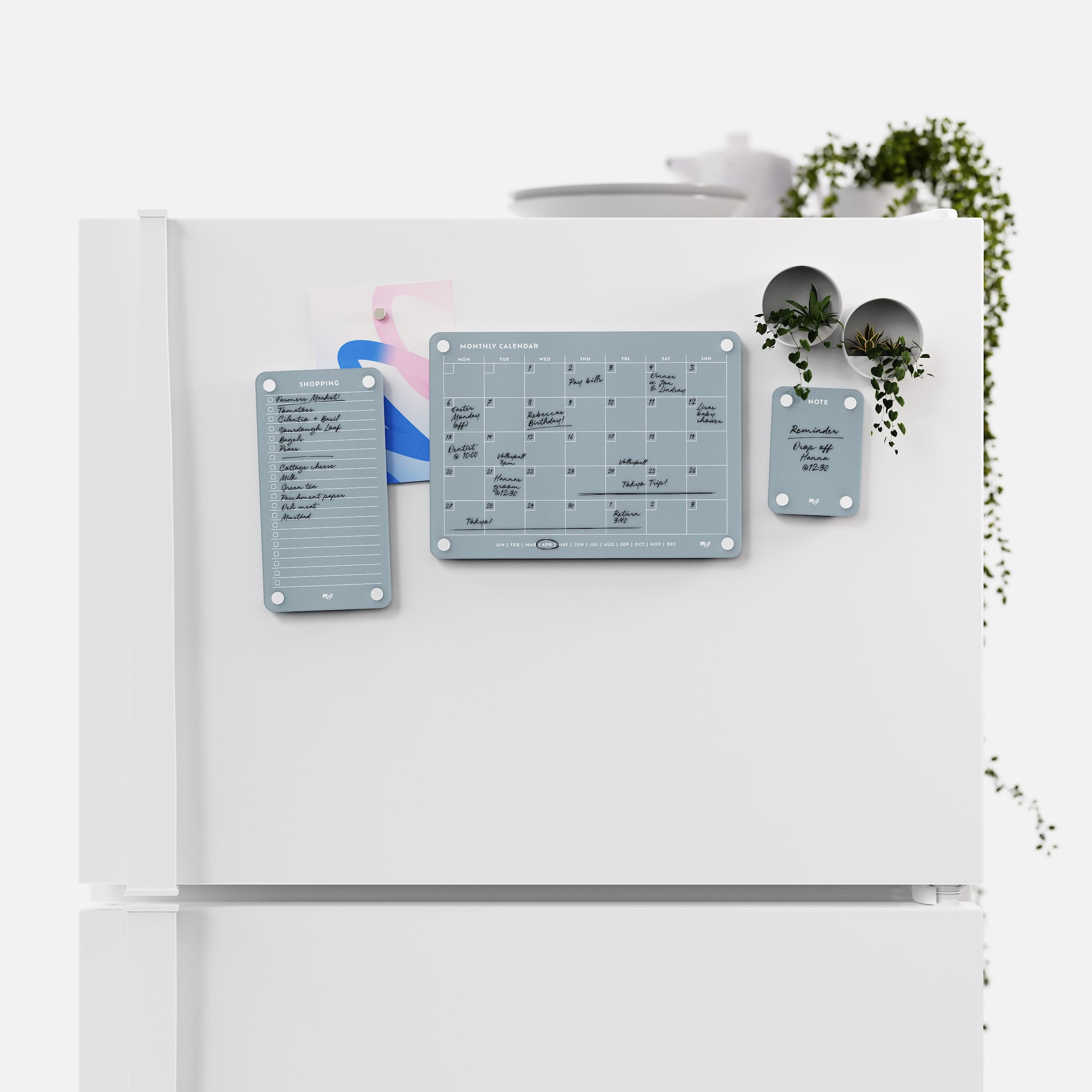 Slate grey shopping list, monthly calendar and note board arranged across a white fridge door